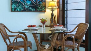 Et rundt glasbord med fletstole, et maleri af et tropisk landskab, en lampe og en vase med blomster.