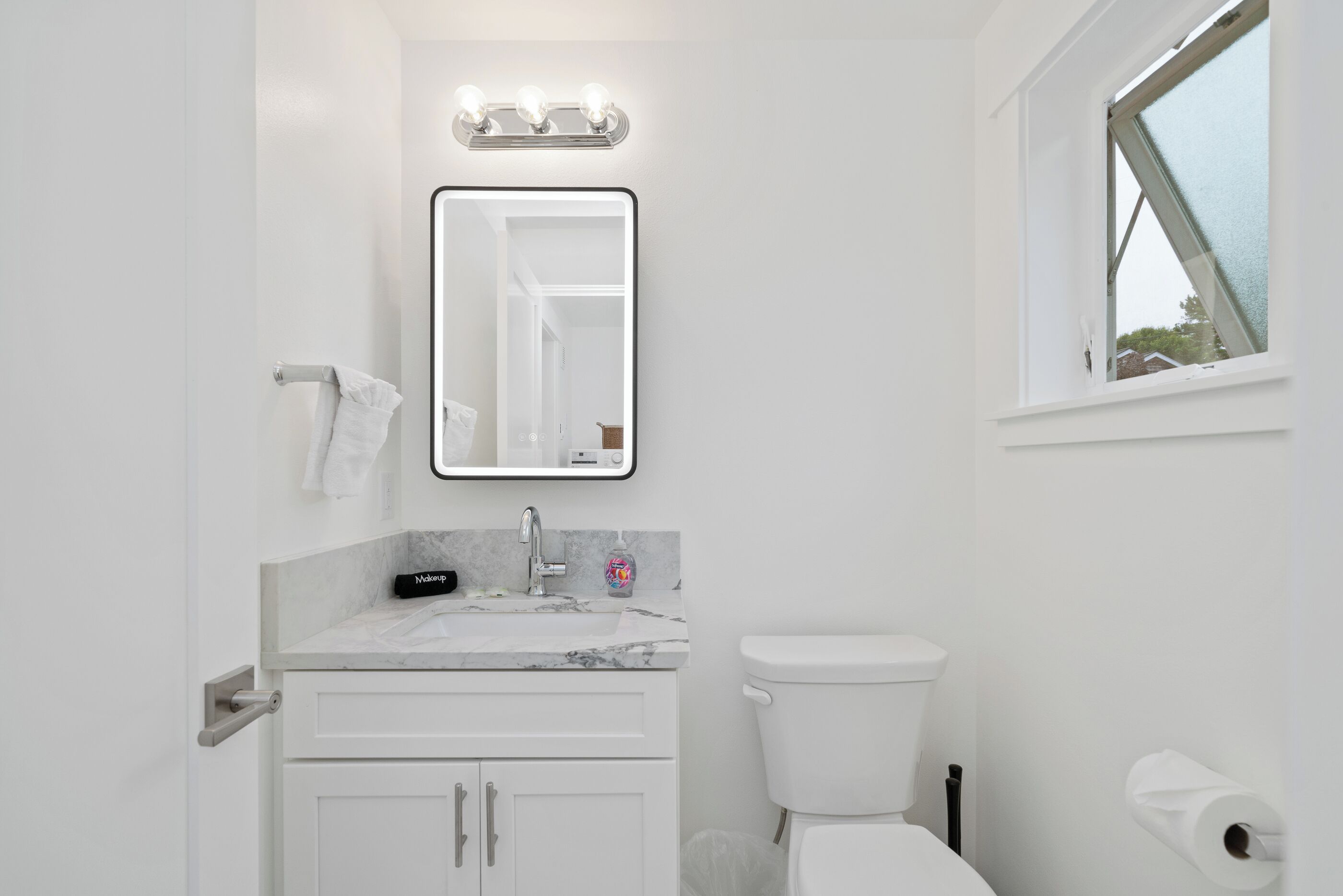 Un baño con lavamanos blanco, un espejo, inodoro y ventana.