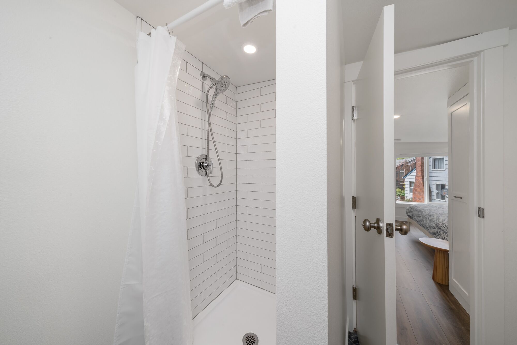 A modern bathroom with white subway tiles, a shower with a handheld showerhead, and a view of the outside through an open door.