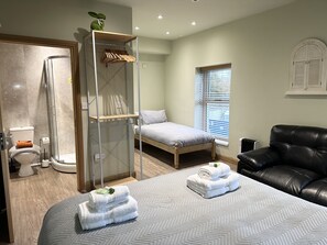 A bedroom with a bed, a black leather sofa, a wooden wardrobe, a shower, and a window with blinds. - Waterfall Country Escape (Neath)