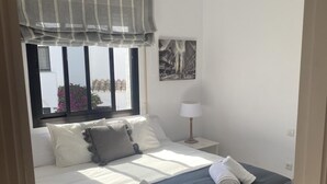 Een netjes opgemaakt bed met twee opgerolde handdoeken, een bedlamp en een raam met uitzicht op gebouwen en bloemen.