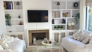 Een moderne woonkamer met een open haard, witte banken, een glazen salontafel en een aan de muur bevestigde televisie.