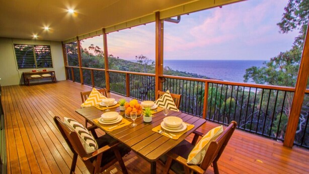 A wooden deck with a table set for dining, overlooking a scenic view of the ocean and trees. - Finding Solace - Sunrise at 1770 (Agnes Water)