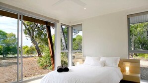 A modern bedroom with a large bed, a wooden headboard, and a view of trees through floor-to-ceiling windows. - Drishti - Sunrise at 1770 (Agnes Water)