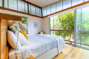 Quarto com uma grande janela, cama com roupa de cama listrada, abajur e um quadro na parede.