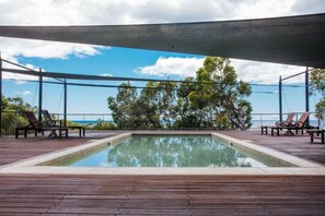 Uma piscina retangular com deck de madeira, rodeada por espreguiçadeiras e um toldo.