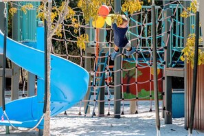 Um parquinho com escorrega azul, rede de escalada e uma árvore.