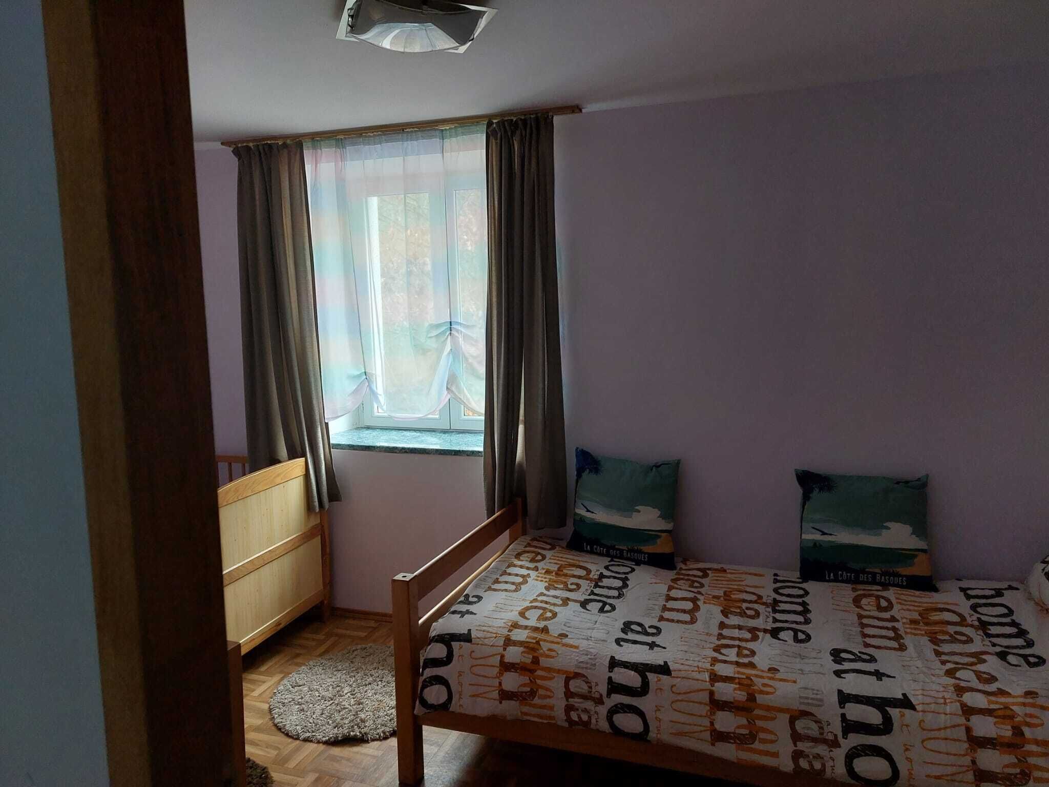 A bedroom with a wooden bed, a window with curtains, a small wooden dresser, and a round rug on the floor.