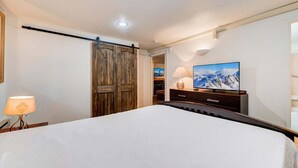 A bedroom with a large bed, a wooden wardrobe, a television on a stand, and a lamp on a bedside table. - Edelweiss Haus G | Hot Tub | Pool | Kitchen | Walk to Slopes (Park City)