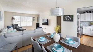 A modern living room with a grey sofa, a dining table with blue plates, and a kitchen area with white cabinets. - Your Up North Getaway! (Traverse City)