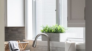 A modern kitchen with a stainless steel sink, a hanging light fixture, and a window with a view of a brick wall and greenery. - Your Up North Getaway! (Traverse City)