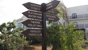 Segnaletica direzionale con diverse frecce che indicano varie strutture come un campo da tennis, un'area giochi, un parco acquatico e un club per bambini.