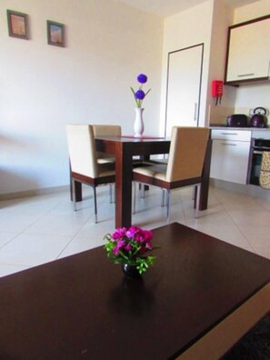 Un coin repas avec une table en bois et quatre chaises, un vase rempli de fleurs violettes, et une cuisine en arrière-plan.