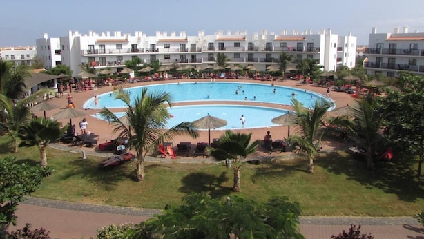 Un resort con una grande piscina circolare circondata da poltrona a sdraio e ombrelloni, con edifici a più piani sullo sfondo.
