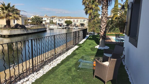 A waterfront patio with a pool, a grassy area, and a white building with a balcony. - Villa Marina , vue sur le Canal, à 5 Minutes du Centre Historique (Aigues-Mortes)