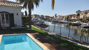 A swimming pool with a wooden deck, a view of a marina with boats, and a white building with a tiled roof. - Villa Marina , vue sur le Canal, à 5 Minutes du Centre Historique (Aigues-Mortes)