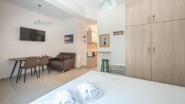 A modern hotel room with a large bed, a desk with chairs, a TV, and a kitchenette. - Cozy Acropolis Studio by Cloudkeys (Athens)