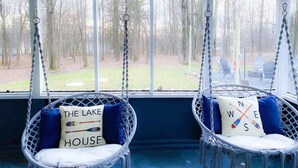 Deux fauteuils en osier, dotés de coussins et accompagnés d’oreillers décorés sur le thème de la marine, sont placés dans un espace abrité avec de grandes fenêtres donnant sur une cour.