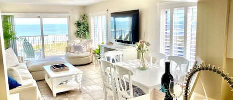 A spacious living area with a balcony overlooking the sea, a flat-screen TV, and a dining table with chairs.