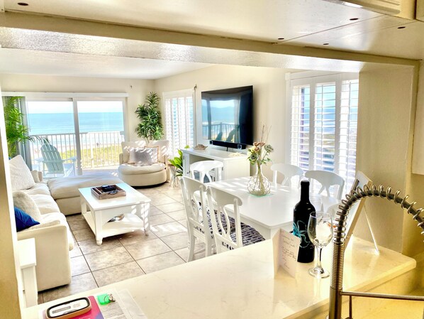 A spacious living area with a balcony overlooking the sea, a flat-screen TV, and a dining table with chairs.