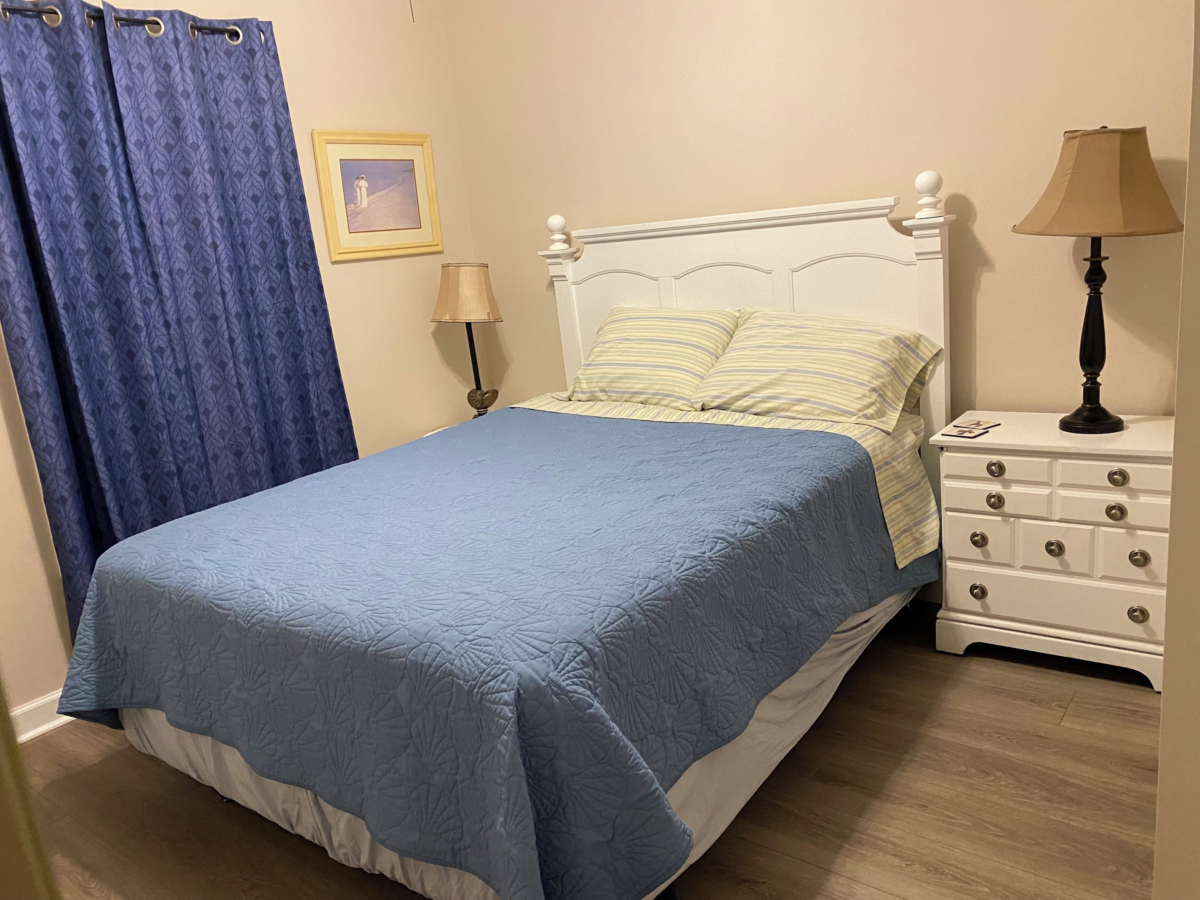 A bedroom with a bed, a nightstand, a lamp, a picture on the wall, and a blue curtain.