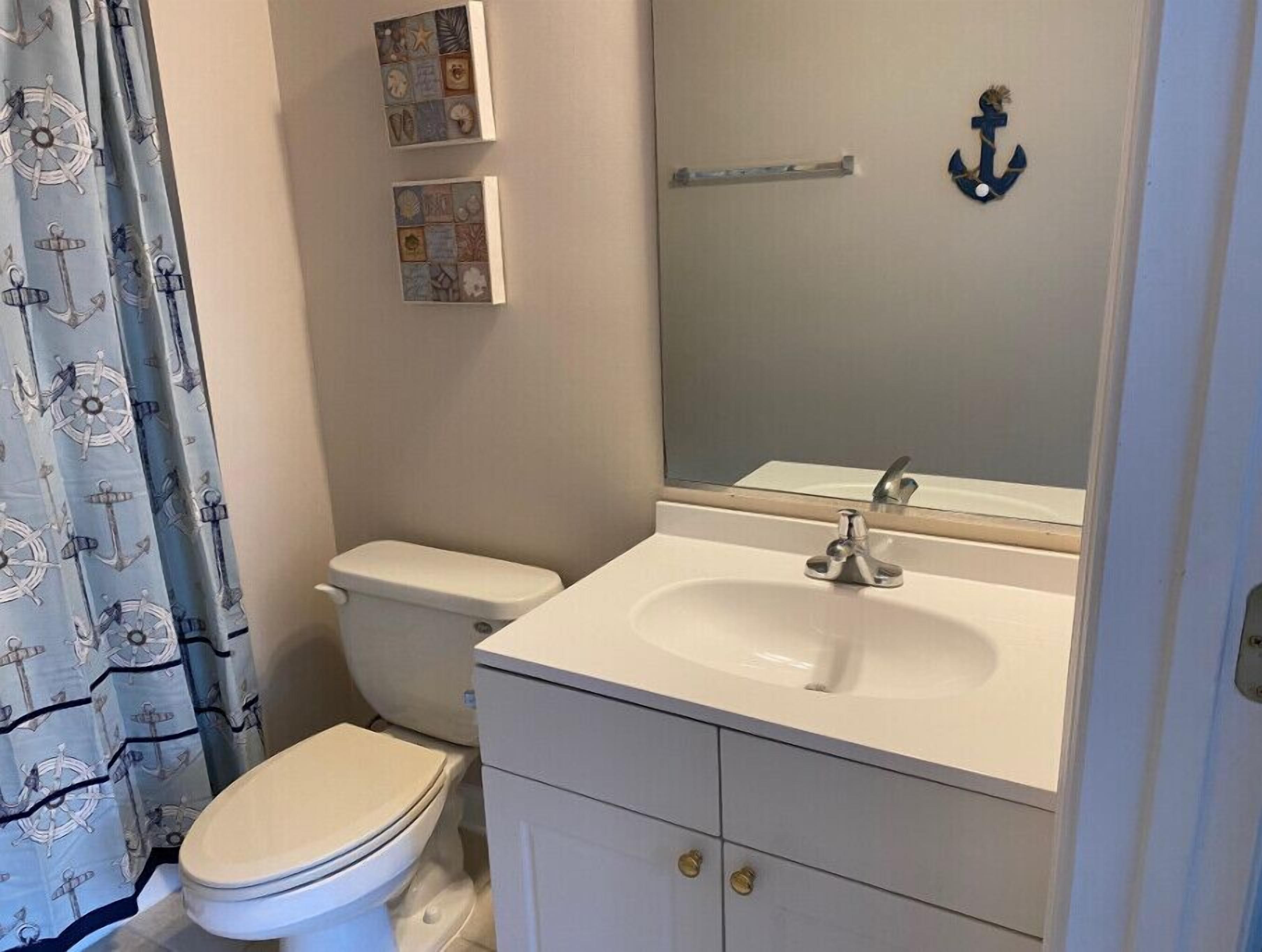 A bathroom with a toilet, sink, and mirror. There are nautical-themed curtains and a small decorative item on the wall.