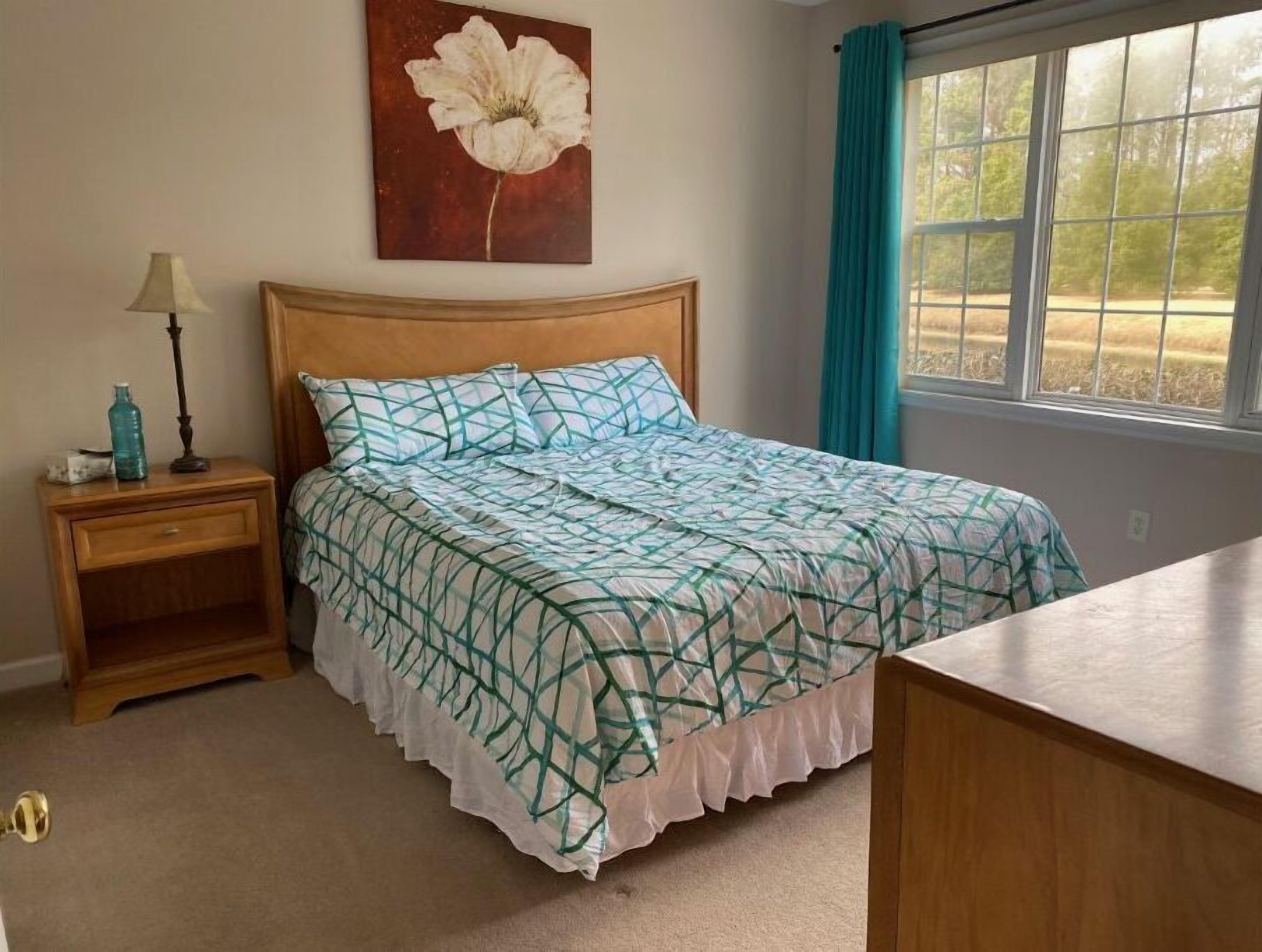 A bedroom with a wooden bed, a nightstand with a lamp, a painting of a flower on the wall, and a window with curtains.
