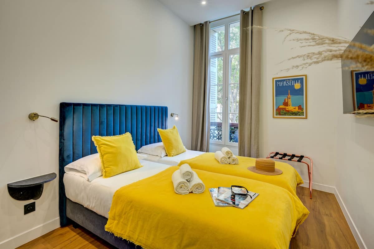 A hotel room with two beds, each with a yellow bedspread and white linens. There is a blue headboard, a small shelf, and a window with curtains.
