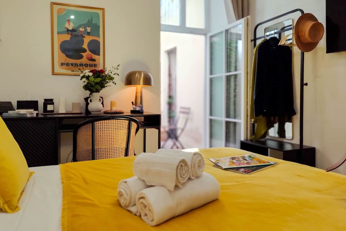 A hotel room with a yellow bedspread, rolled towels, a desk with a lamp, a chair, and a poster on the wall.