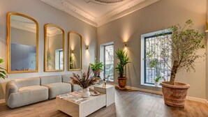 A spacious living room with a grey sofa, a coffee table with magazines, and large mirrors on the wall. There are potted plants and a large window with a view of the city. - Boutique Hôtel Provencia (Marseille)