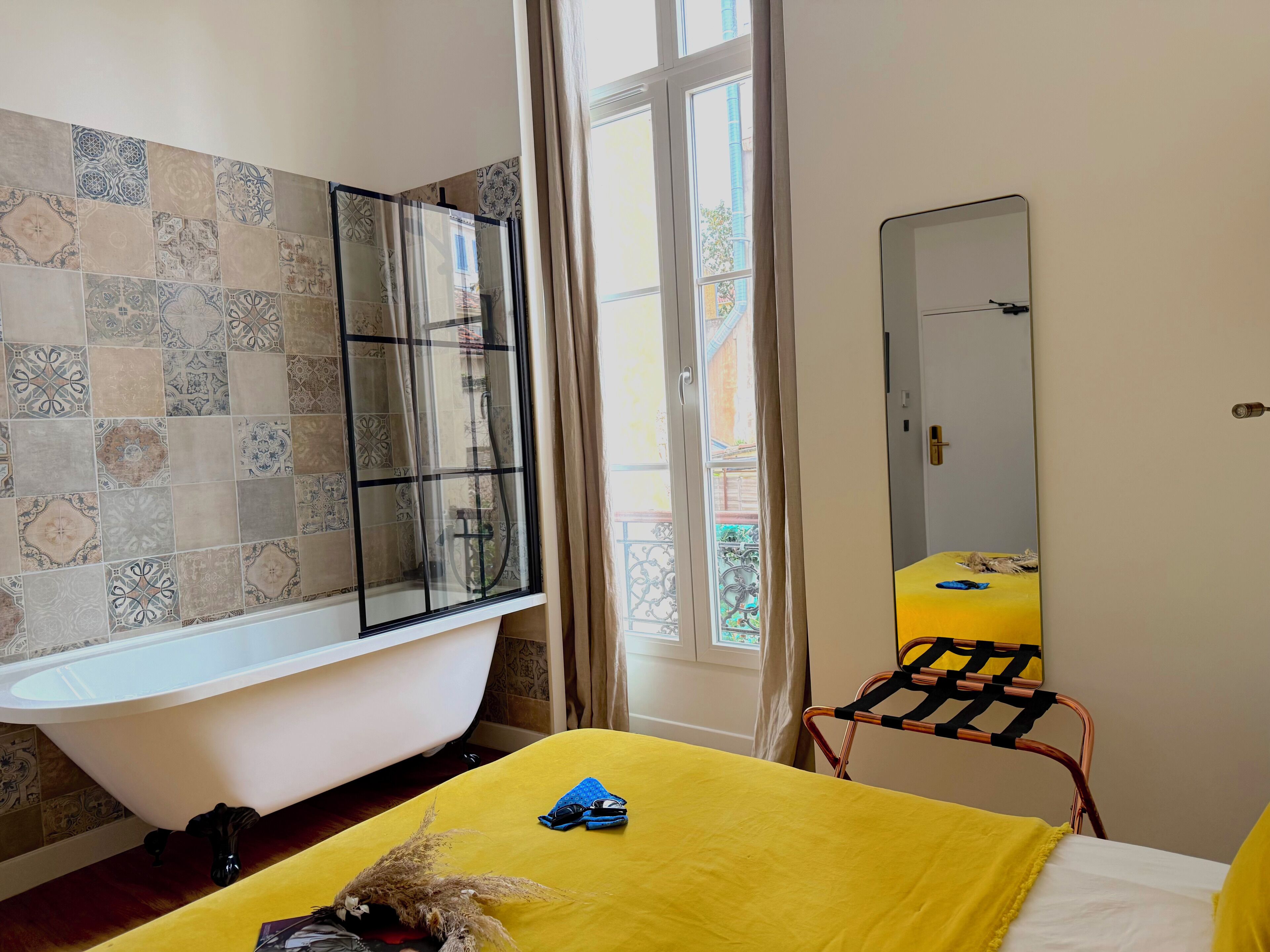 Un baño con bañera, una cama amarilla, un espejo y una ventana con cortinas.