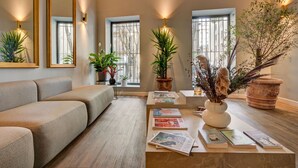 A modern living room with a beige sofa, a coffee table with books and decorative plants, and large windows with natural light. - Boutique Hôtel Provencia (Marseille)