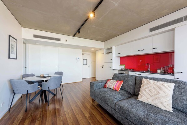 Een moderne woonruimte met een eethoek, een grijze bank met decoratieve kussens, en een keuken met witte kasten en een rode spatwand.