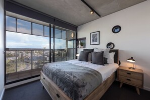 Un dormitorio moderno con una cama grande, una mesita de noche, una lámpara y vistas a la ciudad a través de un amplio ventanal.