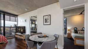 Un interior moderno con una mesa de comedor redonda, sillas grises, un edredón blanco y rojo a cuadros, y un ventanal con vista a la ciudad.