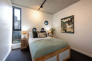 A bedroom with a large window, a bed with a green bedspread, two bedside lamps, and a wall-mounted circular mirror.