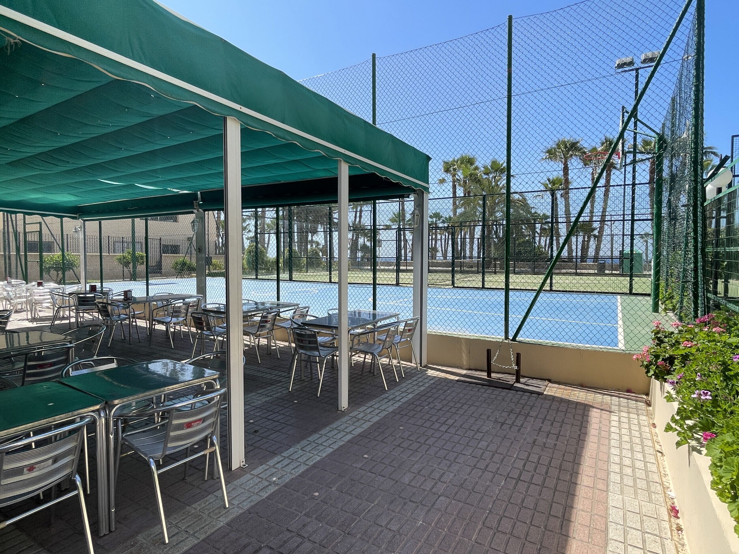 An outdoor seating area with tables and chairs, a covered section, and a view of a tennis court.