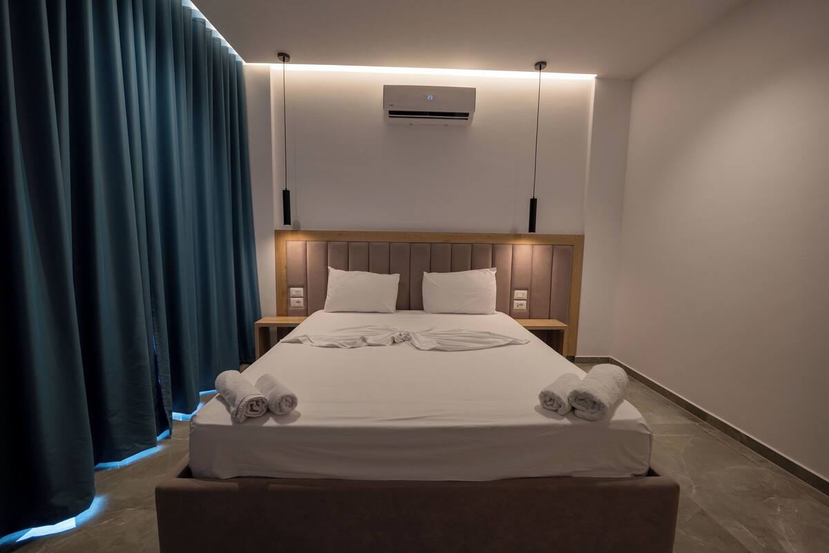 Moderni hotellihuone, jossa on siististi pedattu sänky, integroidulla valaistuksella varustettu pääty, ilmastointilaite ja suuri ikkuna, jossa on verhot.