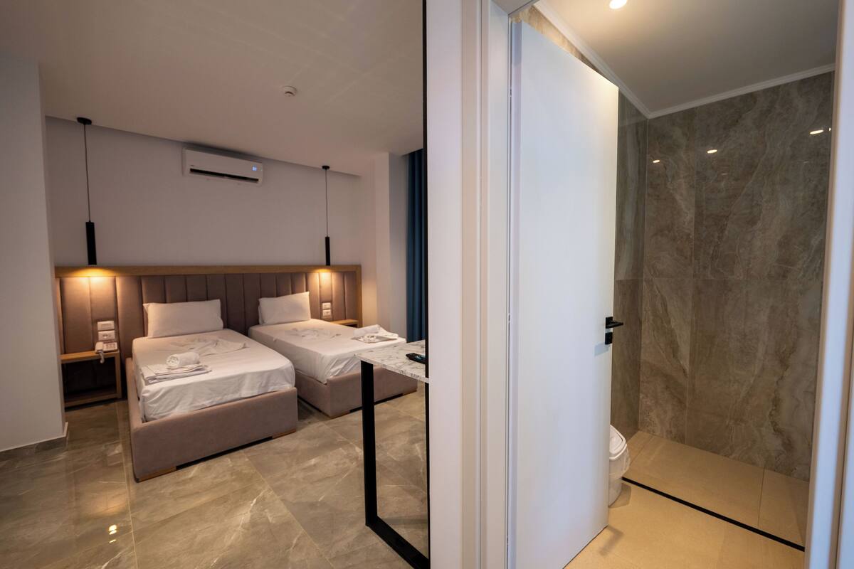 Moderni hotellihuone, jossa on kaksi sänkyä, marmorilla verhoiltu suihku ja lasiseinillä erotettu kylpyhuone.