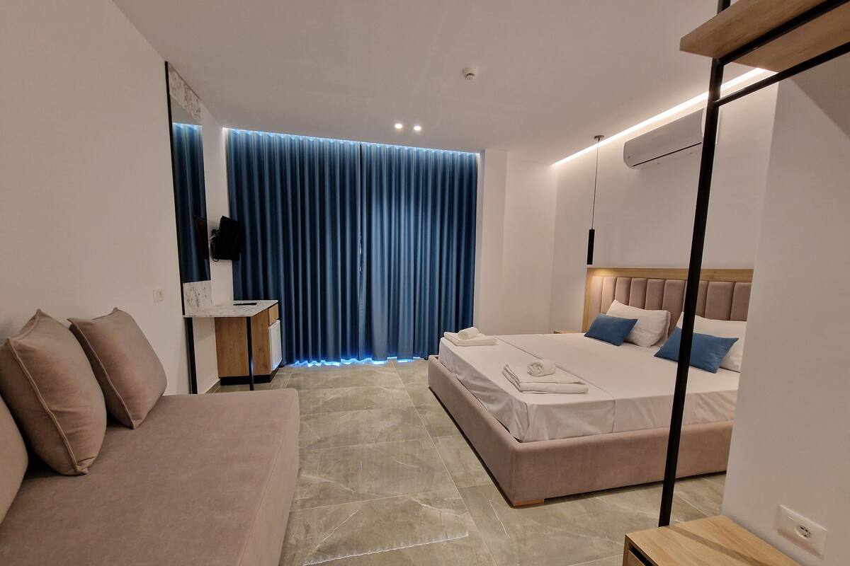 Moderni hotellihuone, jossa on sänky, sohva, yöpöytä ja suuri ikkuna, jossa on verhot.