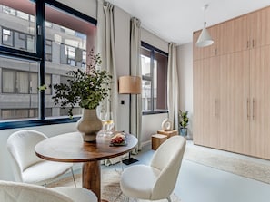 Uma sala de estar moderna com uma mesa de madeira redonda, duas cadeiras brancas, uma janela grande com cortinas, um abajur alto e um guarda-roupa.