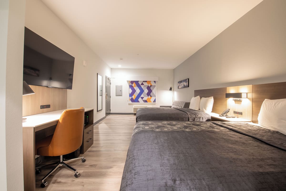 Quarto de hotel com escrivaninha, duas camas, uma cadeira e televisão na parede.
