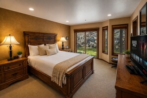 A bedroom with a large bed, wooden headboard, two bedside lamps, a TV, and a view of the outdoors through large windows.