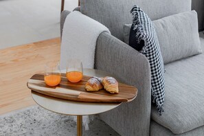 Una bandeja de madera con dos vasos de jugo de naranja y dos croissants sobre una mesita redonda junto a un sofá gris con un cojín de cuadros.