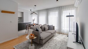 Ett modernt vardagsrum med en grå soffa, ett matbord med stolar, en tv och ett stort fönster med tunna, genomskinliga gardiner.