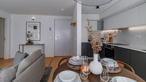 Ett modernt vardagsrum med ett matbord för fyra personer, en grå soffa och ett kök med mörka skåp och en vit bänkskiva.