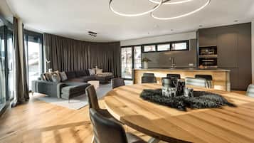 A modern living room with a wooden dining table, dark sofas, and a kitchen area with a large window.