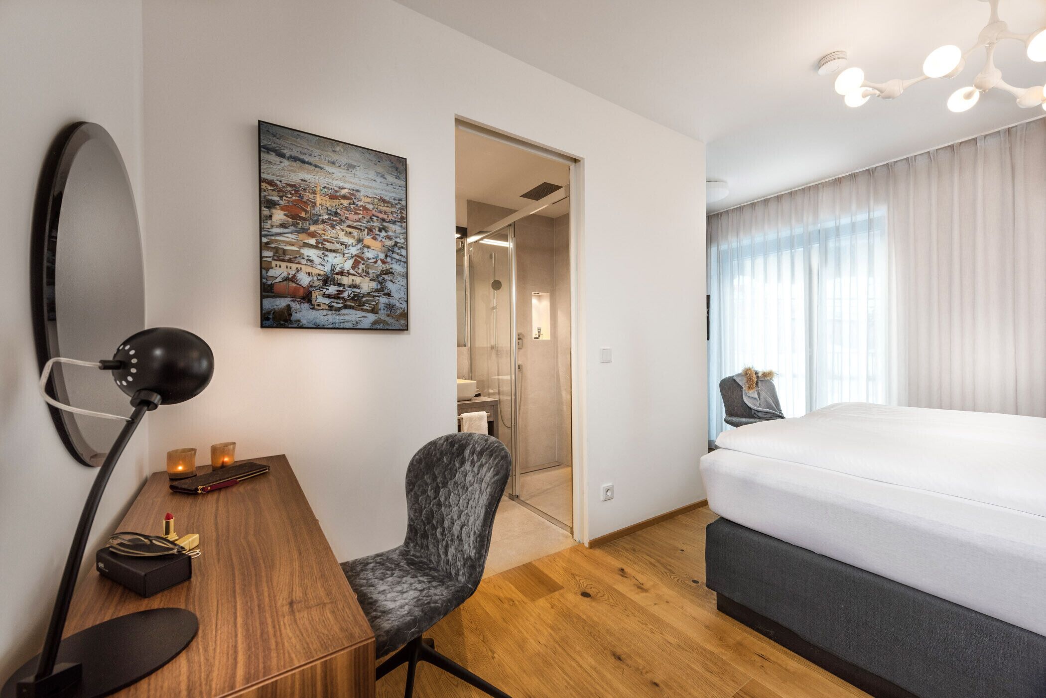 A modern hotel room with a large bed, a desk with a lamp, a chair, a painting, and a shower area.