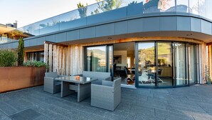 Une maison moderne dotée d’une façade en verre incurvée, d’un espace extérieur aménagé pour s’asseoir et d’un intérieur visible, comprenant une table à manger et des chaises.