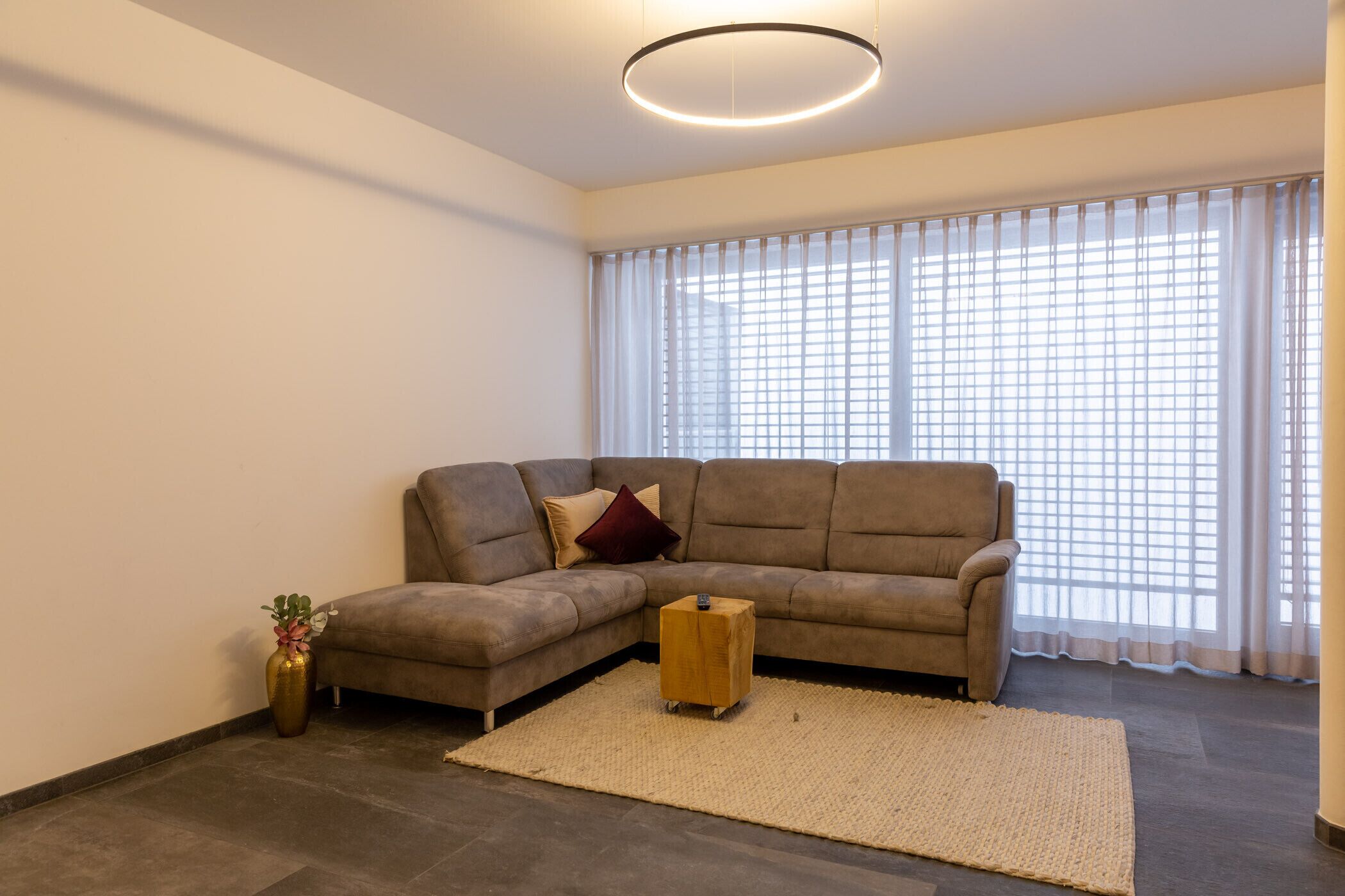 Una sala moderna con un sofá seccional, una pequeña mesita de madera y un ventanal con cortinas de gasa.